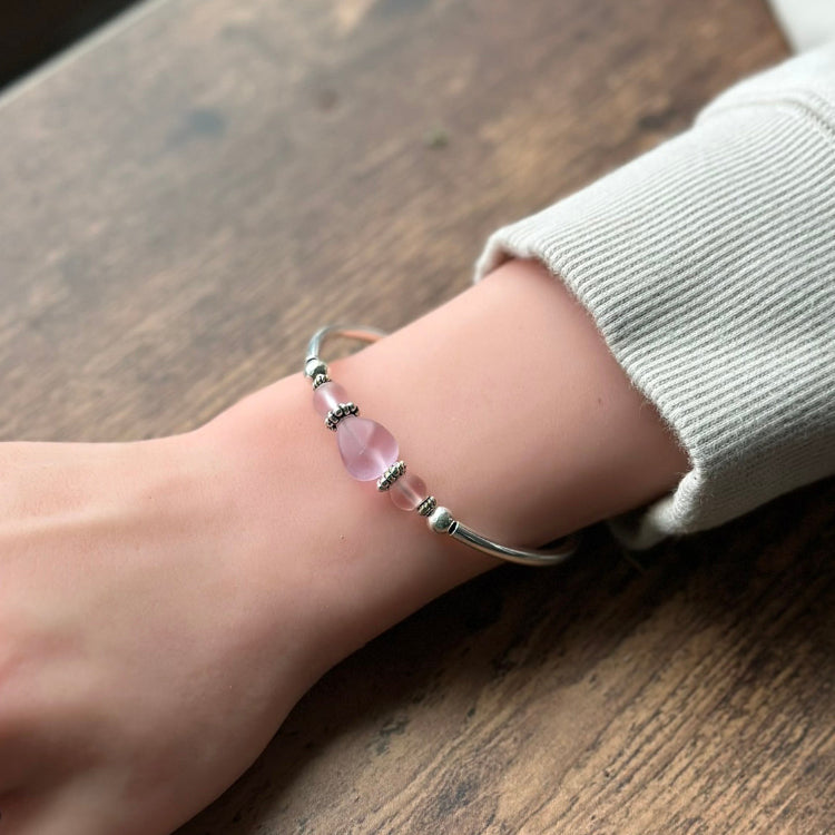 Silver adjustable bangle bracelet worn on the wrist, featuring soft pink frosted sea glass nuggets separated by small decorative silver spacer beads, photographed against a wooden surface.